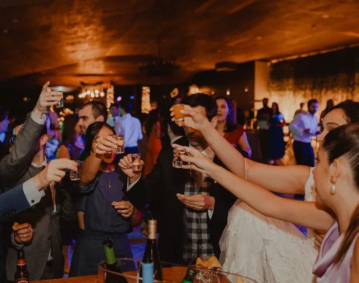 Noiva e convidados a erguer copos de shot num brinde, numa sala de festa à luz quente.